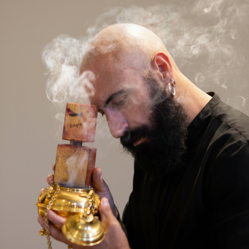 Man holding a smoking incense burner with smoke rising, in a blurred indoor setting.