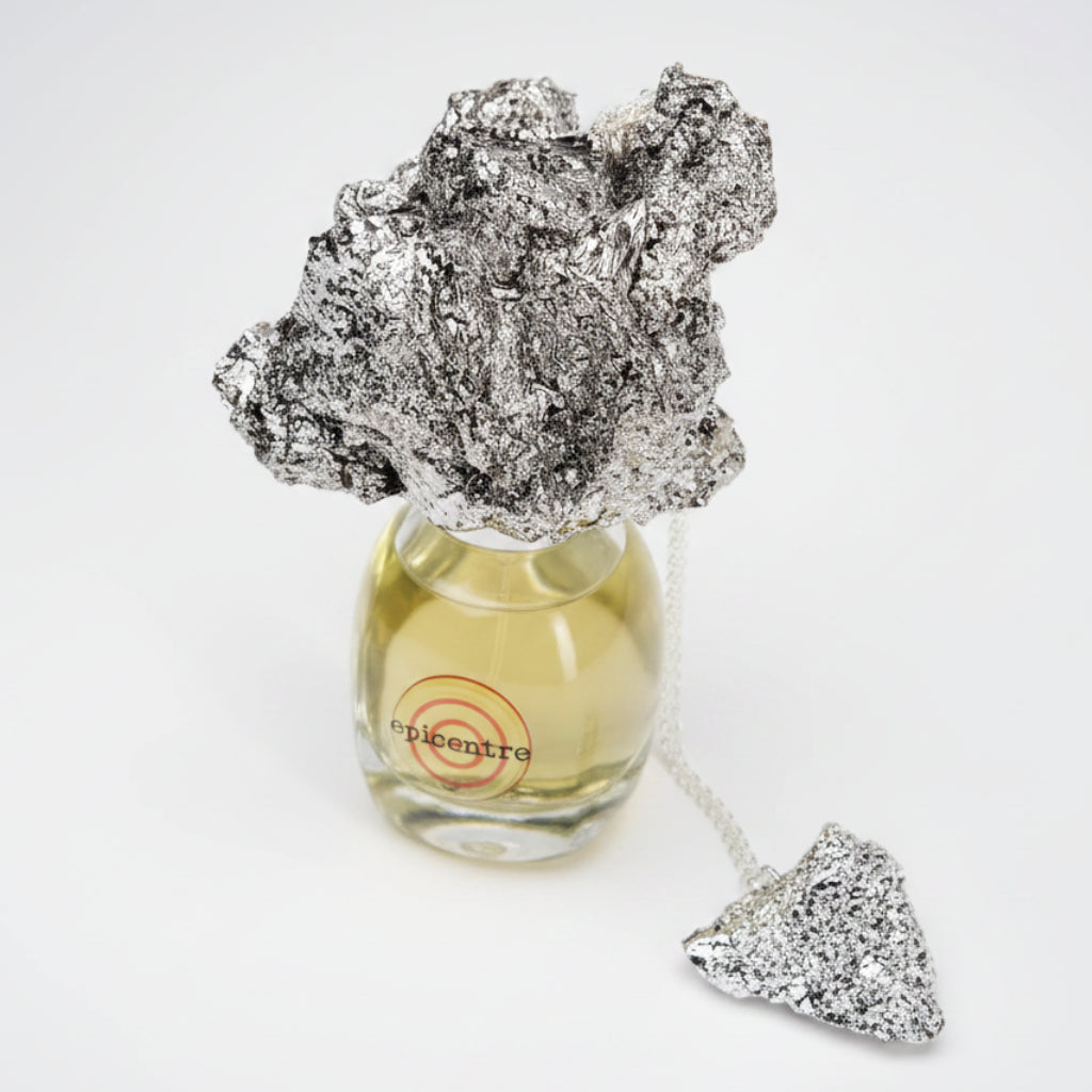 A silver foil rock-shaped perfume atomizer resting on top of a clear glass bottle filled with pale yellow liquid, with a matching foil rock-shaped tag hanging from a chain on a white background.